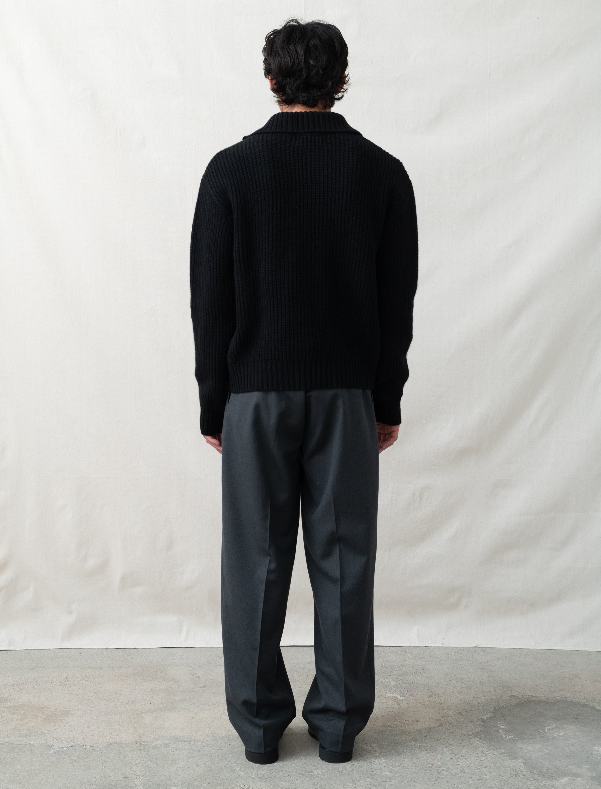 Person wearing a black sweater and gray pants standing against a plain white background